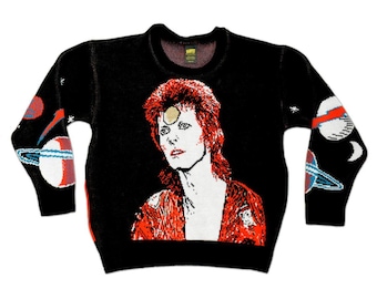 Puede incluir: Un suéter de punto negro de manga larga con un gráfico grande y detallado de David Bowie con cabello rojo y un círculo dorado en la frente. Las mangas están decoradas con motivos de planetas blancos y rojos, y la etiqueta del cuello dice "Bowie".