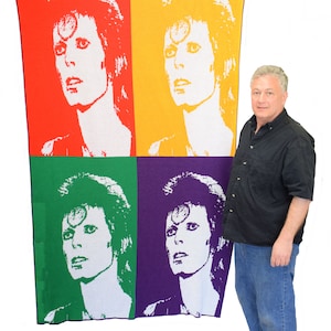 Puede incluir: Una manta de punto con una imagen de estilo pop art de David Bowie con cuatro cuadrados de diferentes colores: rojo, amarillo, verde y morado.