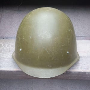 May include: A green military helmet with a worn, textured surface. The helmet has a rounded top and a brim.