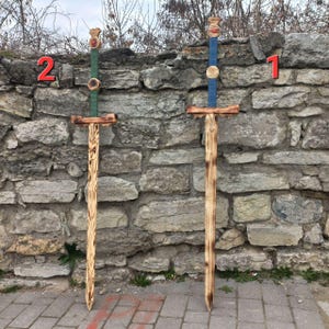 Peut inclure: Deux épées en bois jouet avec des détails peints en bleu et en vert. Les épées sont debout contre un mur en pierre. Le chiffre "1" est écrit sur le mur derrière l'épée bleue et le chiffre "2" est écrit sur le mur derrière l'épée verte.