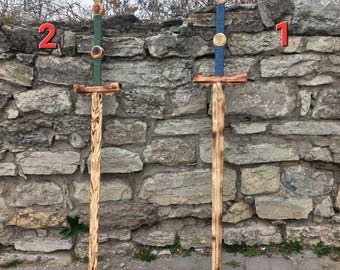 May include: Two wooden toy swords with blue and green painted details. The swords are standing upright against a stone wall. The number "1" is written on the wall behind the blue sword and the number "2" is written on the wall behind the green sword.