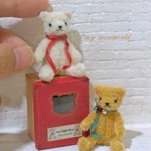 May include: Two small teddy bears, one white and one brown, sitting on a red box with a teddy bear cutout. The box has a label that says "Plushie Teddy Friends".