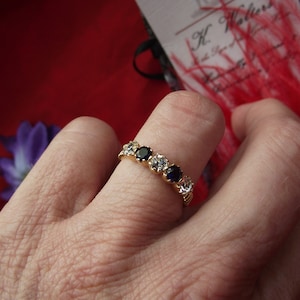 Puede incluir: Un anillo de oro con piedras preciosas transparentes y azul oscuro alternadas. El anillo está en el dedo de una persona.