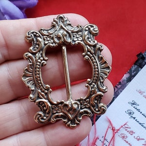 以下が含まれることがあります： 金色の装飾的な金属製のベルトバックル。花柄の細工が施されています。バックルは長方形で、中央に丸みを帯びた楕円形の開口部があります。