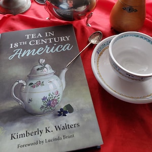 以下が含まれることがあります： 木製の取っ手が付いた銀色のティーポット、18世紀アメリカの紅茶というタイトルの本、ティーカップとソーサー、梨の形をした木製容器、燭台、茶こしを特徴とする静物画。赤い布の上に配置されています。