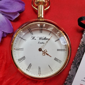 May include: A gold-toned pocket watch with a white face and black Roman numerals. The watch has a clear glass cover and a decorative gold crown. The watch face reads "R. Walters London". The watch is set against a red background.