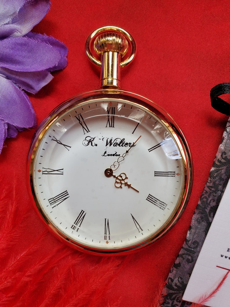 May include: A gold-toned pocket watch with a white face and black Roman numerals. The watch has a clear glass cover and a decorative gold crown. The watch face reads "R. Walters London". The watch is set against a red background.