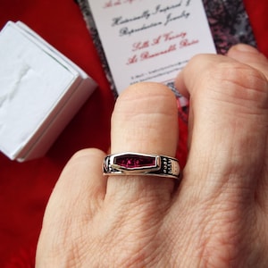 May include: A silver ring with a dark red gemstone set in a rectangular shape. The ring has a textured band with small silver accents.