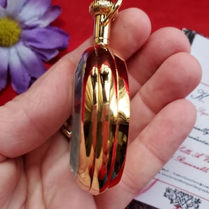 May include: A small, gold-colored perfume bottle with a ribbed design. The bottle has a rounded shape and a decorative top with a small ring. The bottle is held in a hand, with a purple flower and a red background visible.