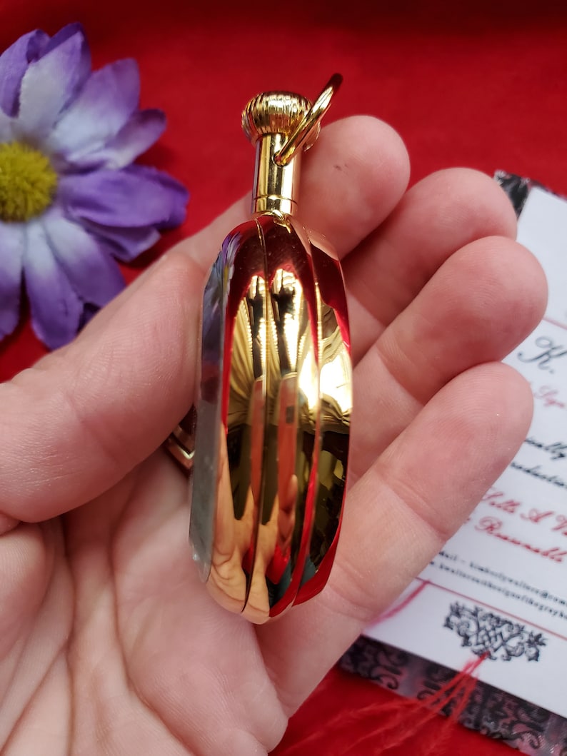 May include: A small, gold-colored perfume bottle with a ribbed design. The bottle has a rounded shape and a decorative top with a small ring. The bottle is held in a hand, with a purple flower and a red background visible.
