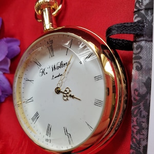 May include: A gold-colored pocket watch with a clear face and Roman numerals. The watch face is white with the text "R. Walters London". The watch is set against a red background.