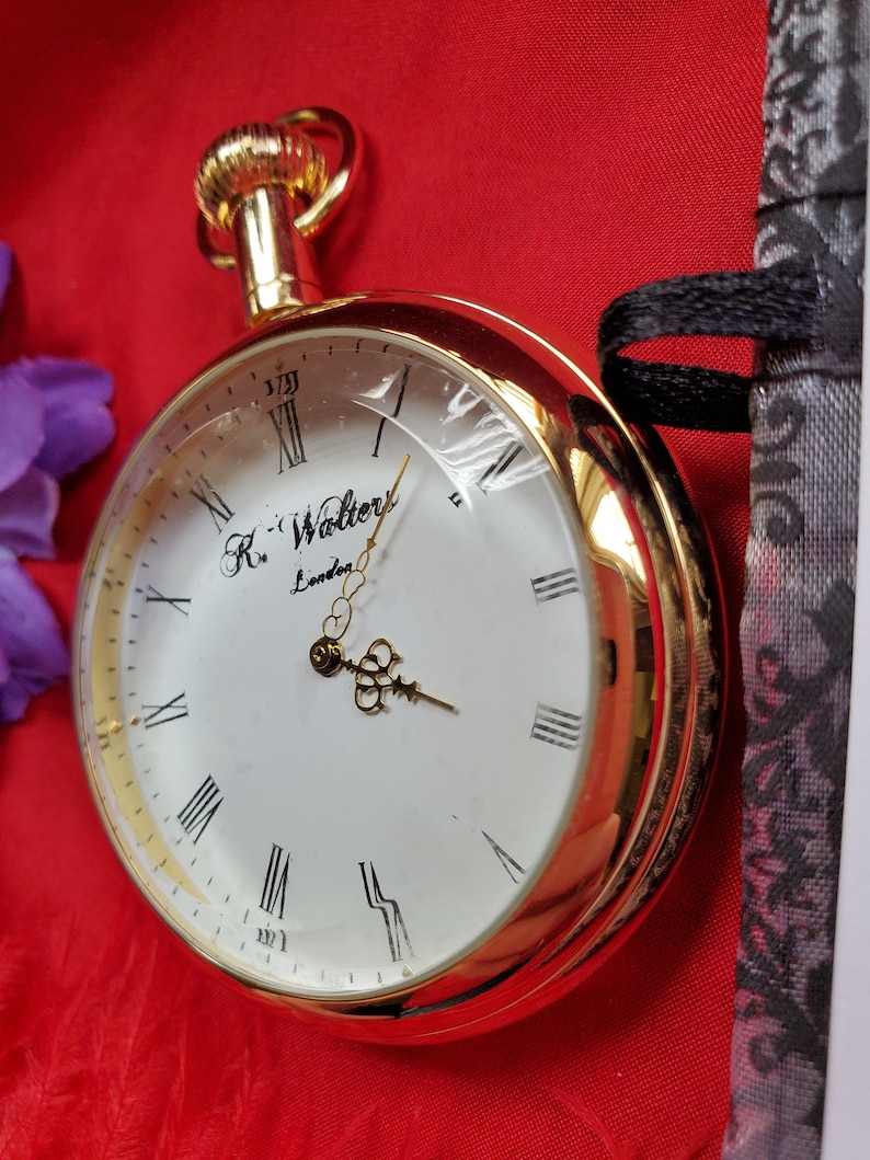May include: A gold-colored pocket watch with a clear face and Roman numerals. The watch face is white with the text "R. Walters London". The watch is set against a red background.