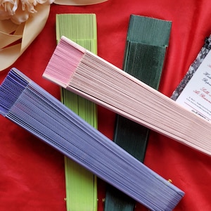 May include: Three folded hand fans in light purple, light green, and dark green, accompanied by a light pink folded fan placed diagonally across them. A white rectangular card with black text reading K. Walters is positioned on the right side.