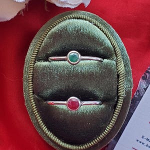 Puede incluir: Dos anillos con piedras preciosas en una caja de anillo de terciopelo verde. El anillo superior tiene una piedra preciosa verde y el anillo inferior tiene una piedra preciosa roja.