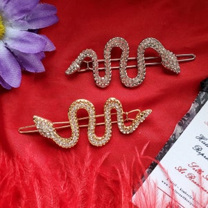 May include: Two gold and silver snake hair clips with rhinestones. The clips are on a red background.