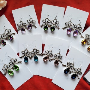May include: A collection of ornate earrings with a bow design and teardrop-shaped gemstones in various colors, including purple, green, and blue. The earrings are displayed on white cards against a red background.