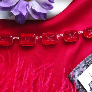 Könnte beinhalten: Eine rote Edelsteinkette mit quadratischen Steinen, verbunden durch silberne Glieder. Die Kette wird auf einem roten Satinhintergrund mit einer lila Blume und rosa Federn präsentiert. Die Edelsteine haben ein eingraviertes Design.