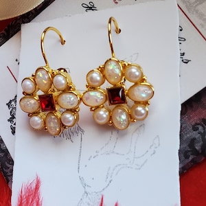 Może przedstawiać: Para złotych kolczyków z czerwonym kamieniem szlachetnym w środku i białymi opalami wokół niego. Kolczyki wiszą na białej kartce z tekstem "W sercu wszystkiego."