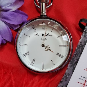 May include: A silver pocket watch with a white face, Roman numerals, and black hands. The watch has the text "K. Walters London" on the face. The watch is on a red background with a purple flower and a decorative card.