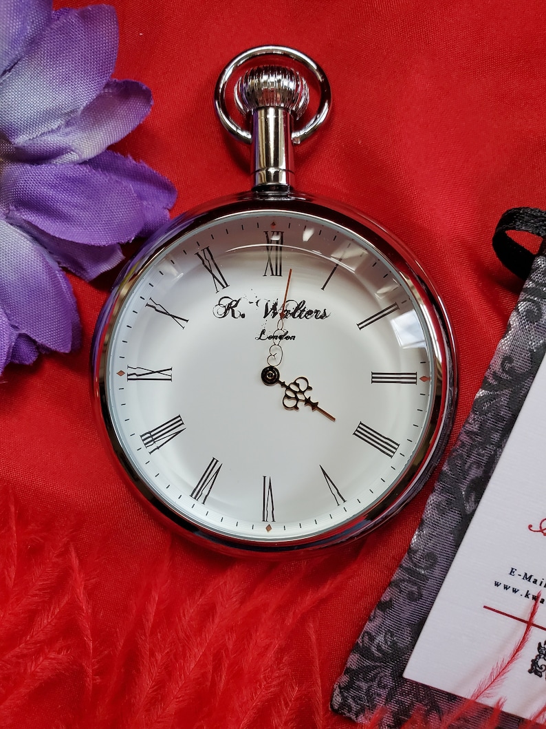 May include: A silver pocket watch with a white face, Roman numerals, and black hands. The watch has the text "K. Walters London" on the face. The watch is on a red background with a purple flower and a decorative card.