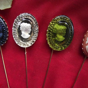 May include: Four decorative pins with different colored glass tops. Each pin features a cameo-style design of a woman's profile. The pins are on a red fabric background.