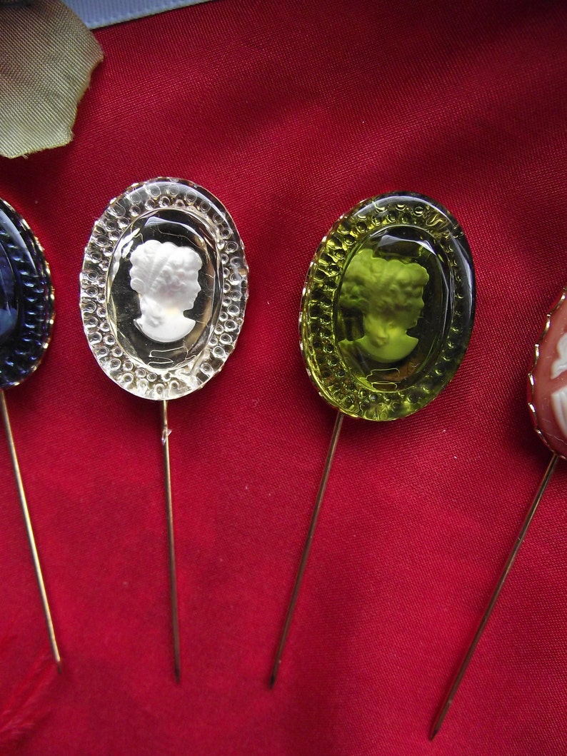 May include: Four decorative pins with different colored glass tops. Each pin features a cameo-style design of a woman's profile. The pins are on a red fabric background.
