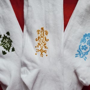 May include: Three white socks with decorative patterns. One sock features a green floral design, another has a gold pattern, and the third displays a blue floral design. The socks are displayed on a red surface.