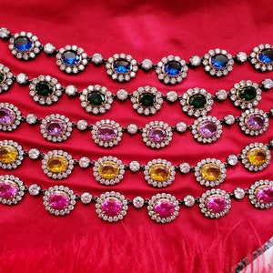 May include: A close-up of six rows of oval-shaped jeweled necklaces. The necklaces are layered on top of each other and feature different colored stones, including blue, green, purple, yellow, and pink.