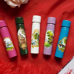 Puede incluir: Cinco estuches cilíndricos de madera en rosa, verde oscuro, blanco, lavanda y azul, cada uno con un paisaje ovalado pintado a mano y enmarcado en oro. Los estuches están dispuestos verticalmente y se ve una esquina de un documento con texto.