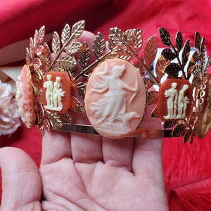 May include: A gold-toned metal crown featuring a series of oval-shaped cameos in shades of peach and terracotta. The crown is decorated with metallic leaf motifs and displays raised white relief figures of mythological scenes on each cameo.