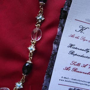 May include: A gold chain necklace with alternating clear glass beads, black faceted beads, and white flower charms with blue centers. The necklace is on a red background with a white card that reads "K. At the Sign of Historically Reproductio Sells A At Reasonabe E-Mail - kimberlywalters@ www.kwaltersatthesignofthegr"
