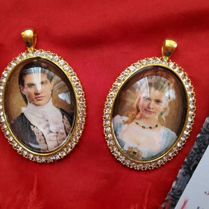 May include: Two gold oval-shaped pendants with clear glass fronts and a white rhinestone border. The pendants feature images of a man and a woman in period clothing.