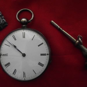 May include: A silver pocket watch with a white face and black Roman numerals, next to a silver watch key with a hoof design, set against a red background. The watch has a loop at the top.