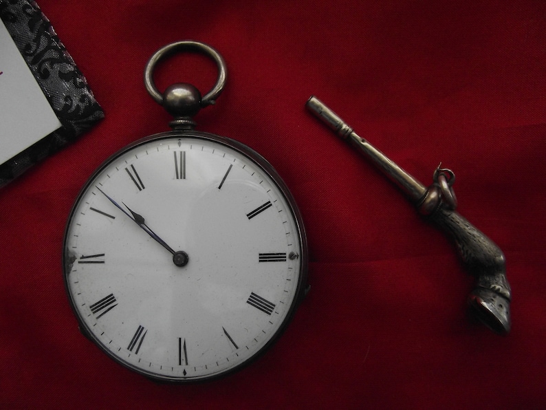 May include: A silver pocket watch with a white face and black Roman numerals, next to a silver watch key with a hoof design, set against a red background. The watch has a loop at the top.