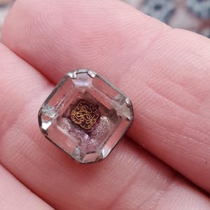 Puede incluir: Un botón de cristal transparente con un diseño dorado en el interior. El diseño es una letra "S" estilizada con una corona encima.