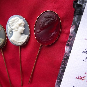 May include: Three decorative pins with a cameo design. The pins are gold-toned and feature a white cameo, a red cameo, and a blue cameo. The cameos are all of a woman's face.