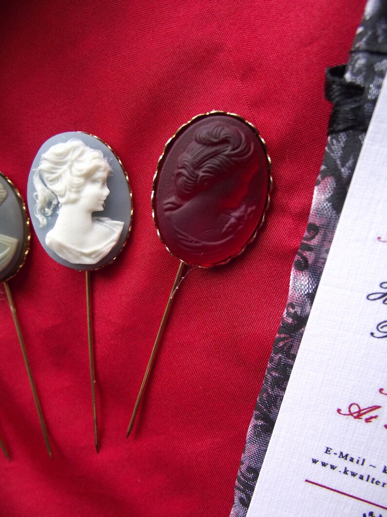 May include: Three decorative pins with a cameo design. The pins are gold-toned and feature a white cameo, a red cameo, and a blue cameo. The cameos are all of a woman's face.
