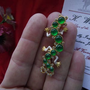May include: A gold-tone lizard brooch with green gem stones and white enamel accents. The brooch is on a hand.