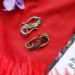 May include: Two ornate metal clasps with floral designs. The clasp on the left is silver and the clasp on the right is brass.