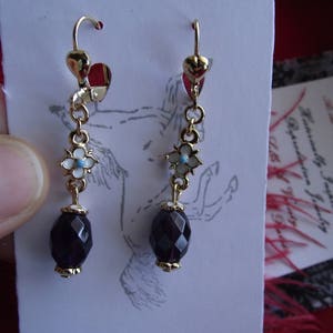 May include: A pair of gold dangle earrings with a dark purple faceted gemstone and white flower accents. The earrings have a heart-shaped hook.