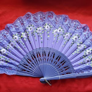 May include: A hand-painted, purple folding fan with a floral design. The fan features a delicate lace trim in a matching purple hue. The fan's wooden ribs are visible at the base, and the handle is a solid purple color.