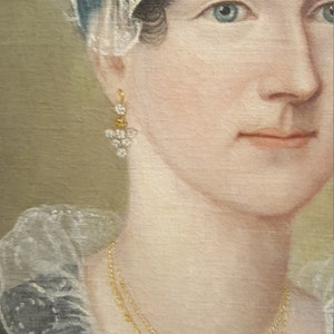May include: A portrait painting of a woman wearing a white lace bonnet with a blue ribbon, a gold necklace, and gold earrings with white stones. The woman has dark hair and blue eyes.