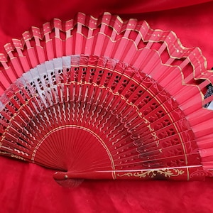 May include: A vibrant red hand fan with intricate cut-out designs and gold accents. The fan is open, displaying its full form against a red fabric backdrop. The fan's wooden frame is visible, and the design is symmetrical.