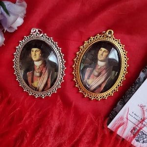 May include: Two silver and gold framed pendants with portraits of men in period clothing. The pendants are on a red background.