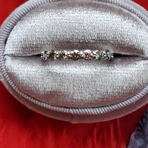 May include: A delicate silver band ring with a row of small, round, multi-colored gemstones. The ring is in a gray velvet ring box.