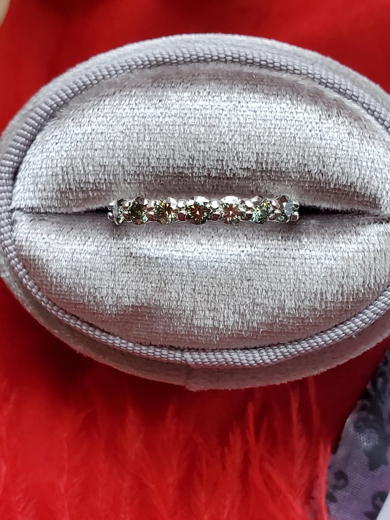 May include: A delicate silver band ring with a row of small, round, multi-colored gemstones. The ring is in a gray velvet ring box.