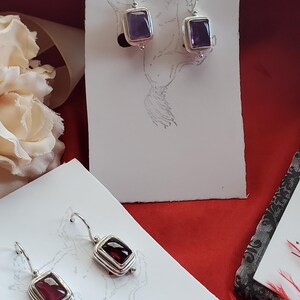May include: Two pairs of sterling silver earrings with square-shaped gemstones. One pair features a purple amethyst gemstone, and the other pair features a red garnet gemstone.