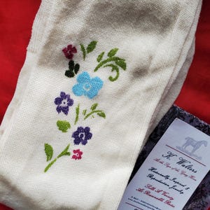 May include: Off-white knit stockings featuring an embroidered floral pattern with blue, purple, and pink flowers and green leaves. A rectangular white business card is included, displaying the text K. Walters, At the Sign of the Gray Horse.