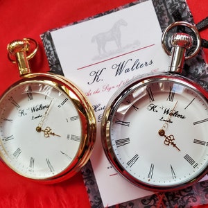 May include: Two pocket watches with white faces and Roman numerals. One watch has a gold-tone case, the other is silver-tone. The watches are displayed on a red surface with a card that reads "K. Walters London."
