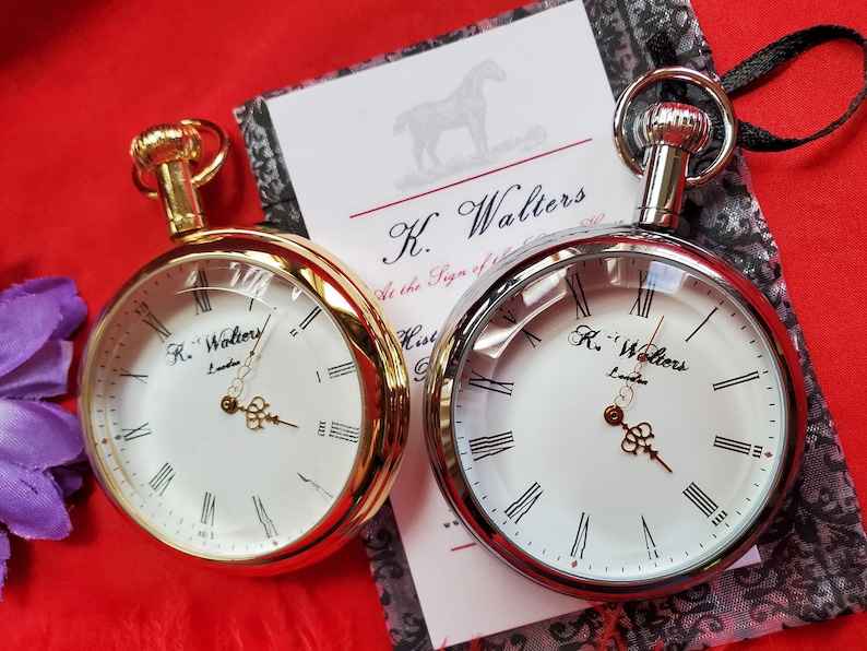 May include: Two pocket watches with white faces and Roman numerals. One watch has a gold-tone case, the other is silver-tone. The watches are displayed on a red surface with a card that reads "K. Walters London."